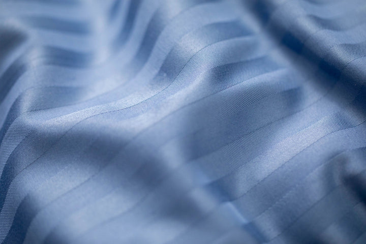 Сатинова тканина насиченого синього кольору з ніжним смугастим візерунком, м’яка та гладка на дотик.