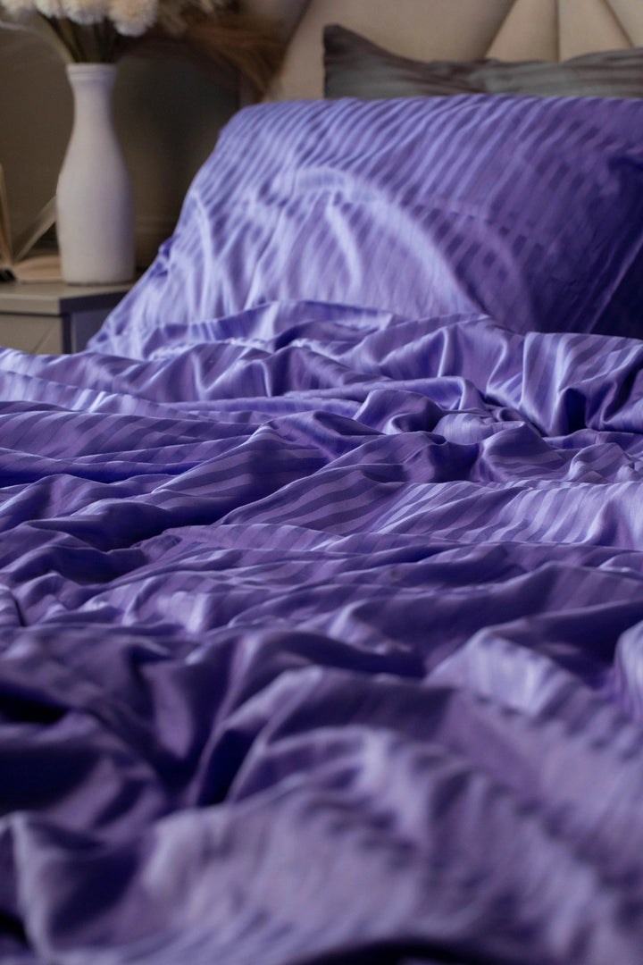 Розкішна смугаста сатинова підковдра насиченого фіолетового кольору, елегантно спущена на ліжко.