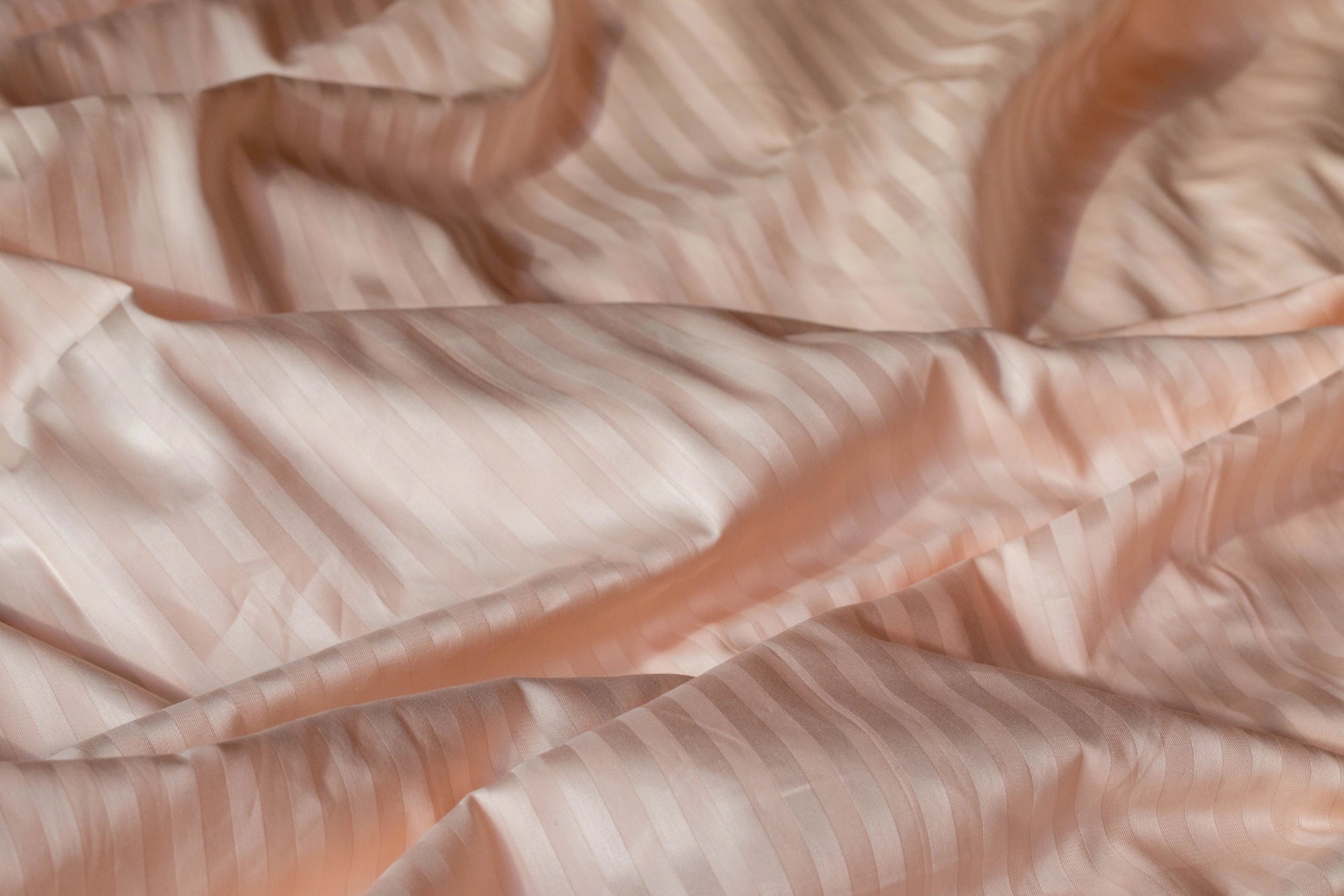 Крупний план персикової сатинової тканини зі смугастим візерунком, що м’яко виблискує на світлі.