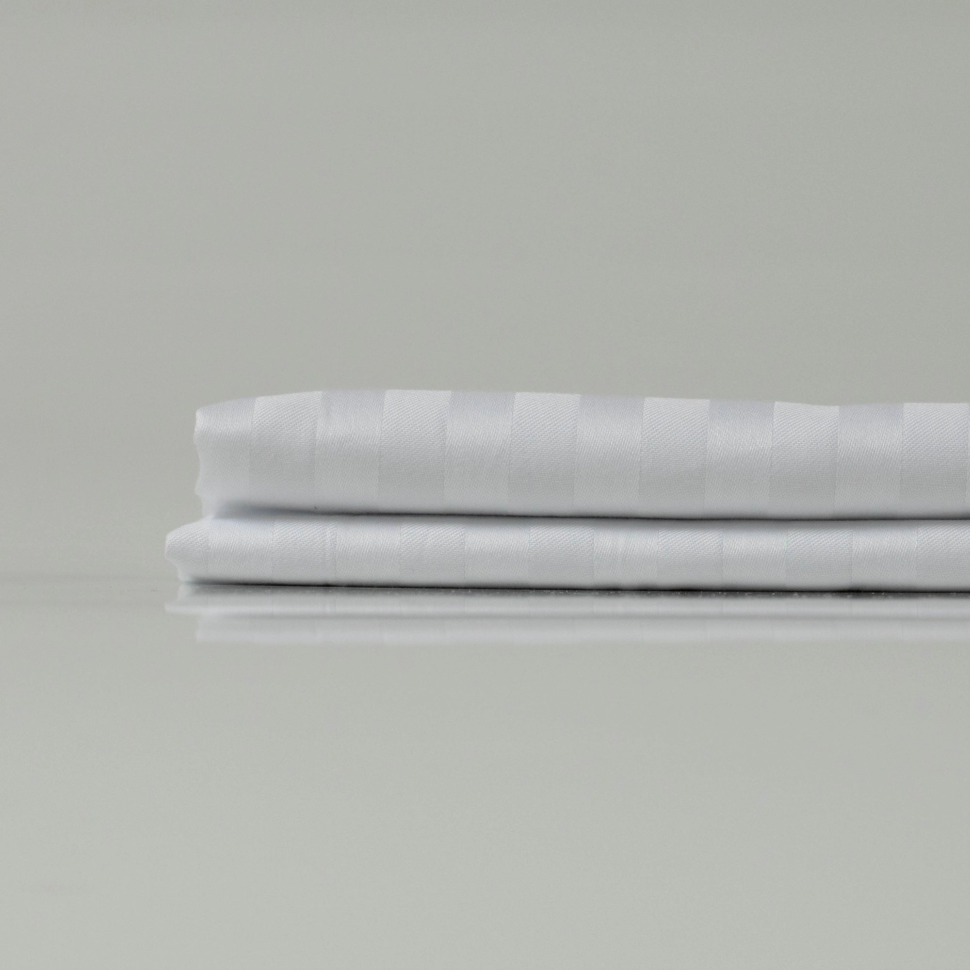 Складене біле простирадло зі страйп-сатину - гладка текстура, ледь помітний блиск і класичний візерунок для елегантної спальні.