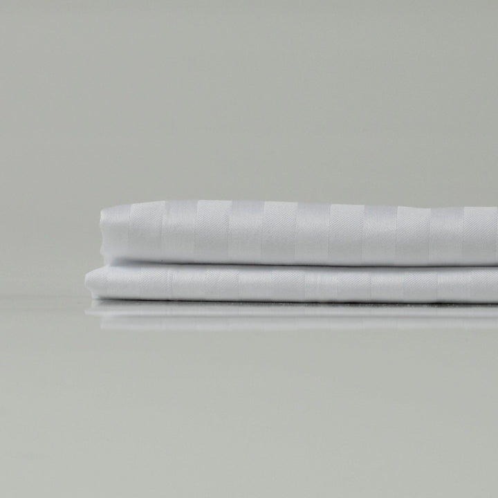 Складене біле простирадло зі страйп-сатину - гладка текстура, ледь помітний блиск і класичний візерунок для елегантної спальні.